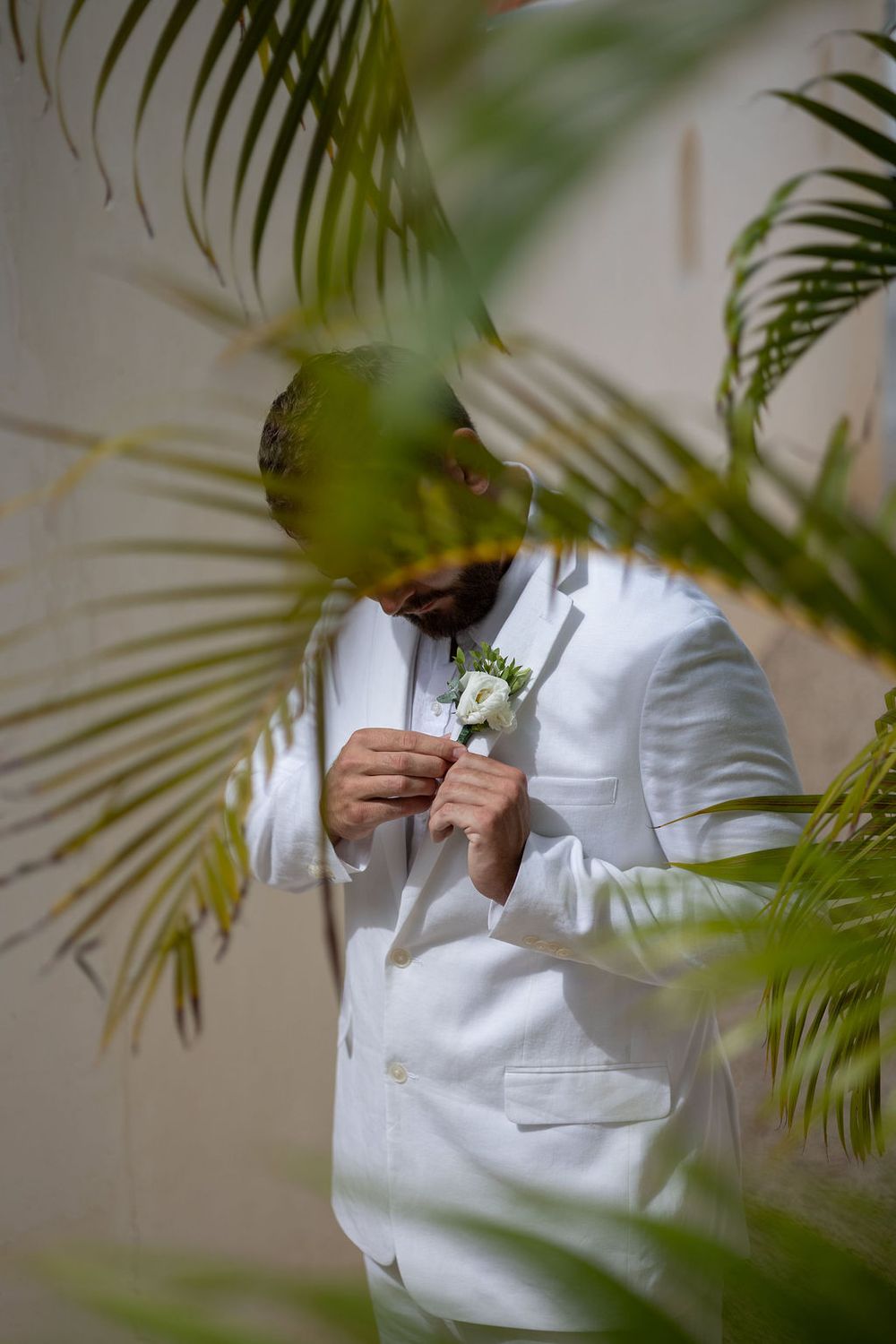 Groom in white suit adjusting boutonniere surrounded by tropical palm leaves at outdoor wedding.