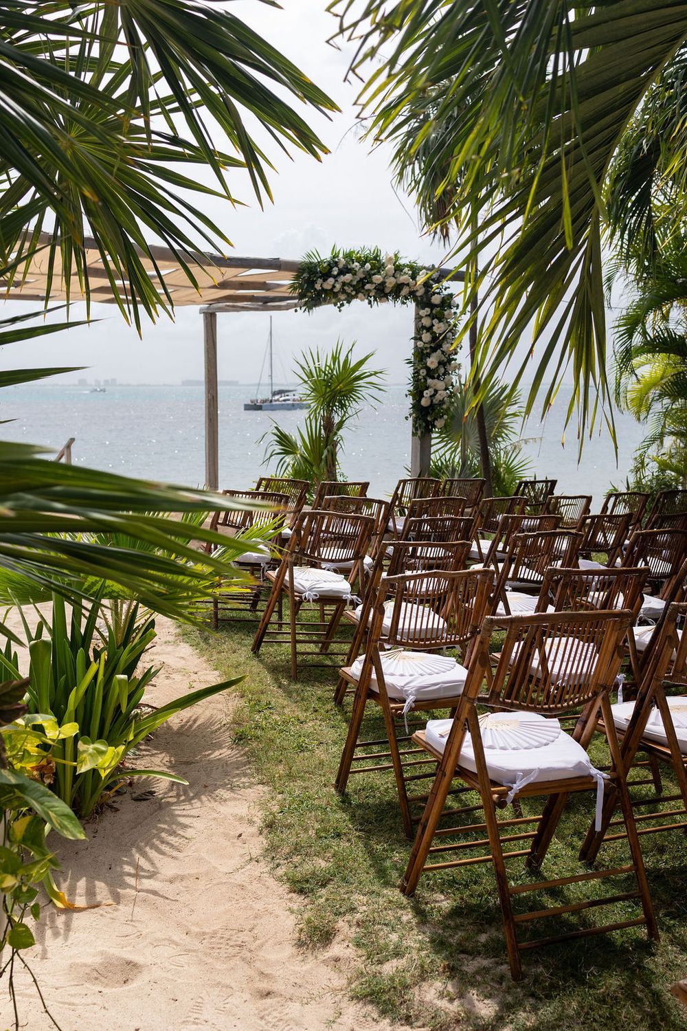 Outdoor beach wedding ceremony setup with wooden folding chairs, floral arch, palm trees, and ocean view with sailboat.