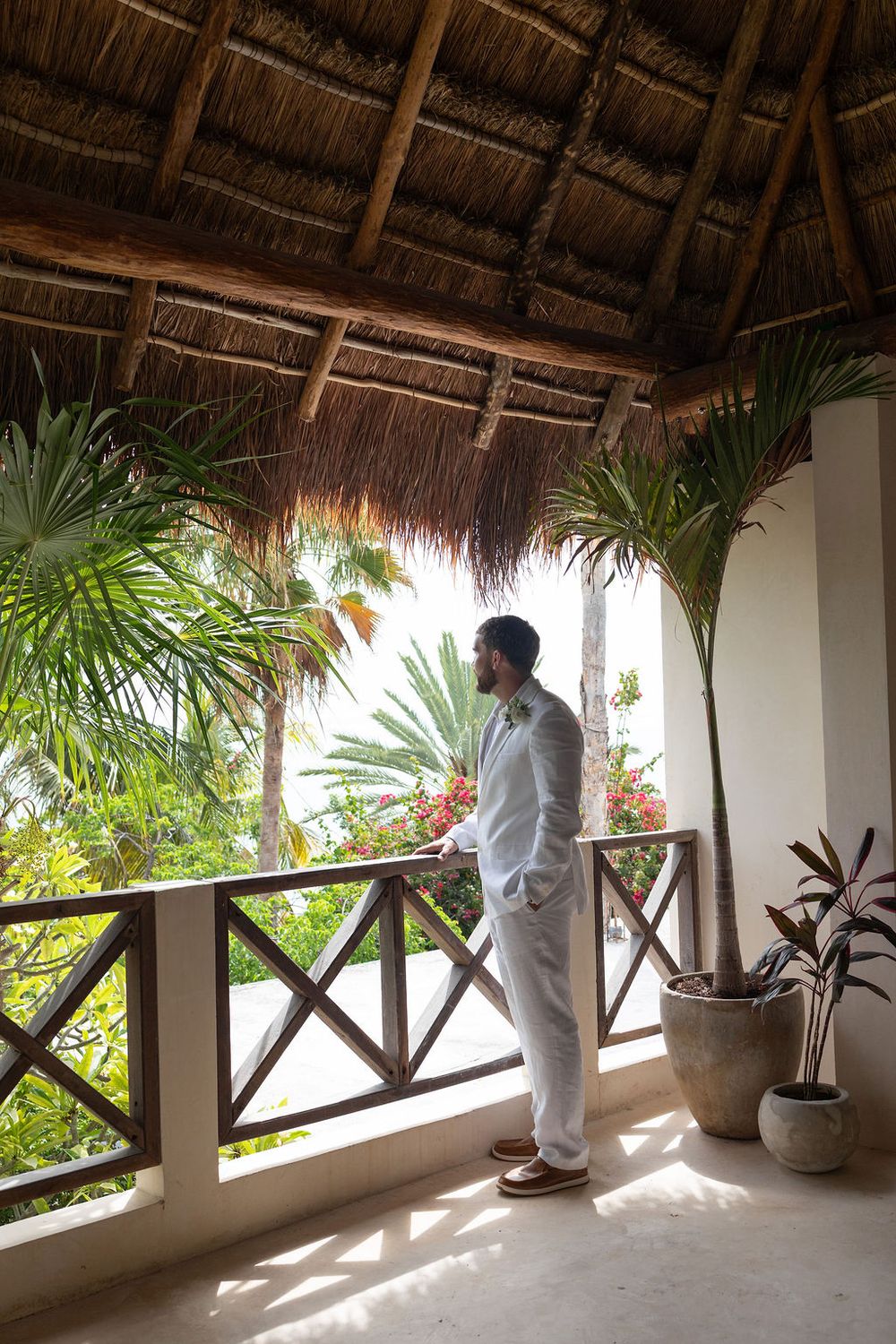Groom in light gray suit stands on tropical resort balcony with thatched roof, surrounded by lush palm trees.