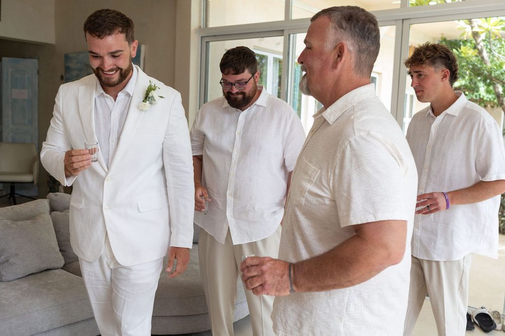 Groomsmen in white suits laughing and getting ready together before a wedding ceremony.