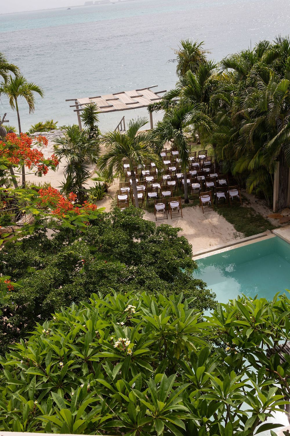 Aerial view of a tropical beachfront resort with pool, palm trees, and rows of chairs set up for an outdoor ceremony at Coco B, Isla Mujeres