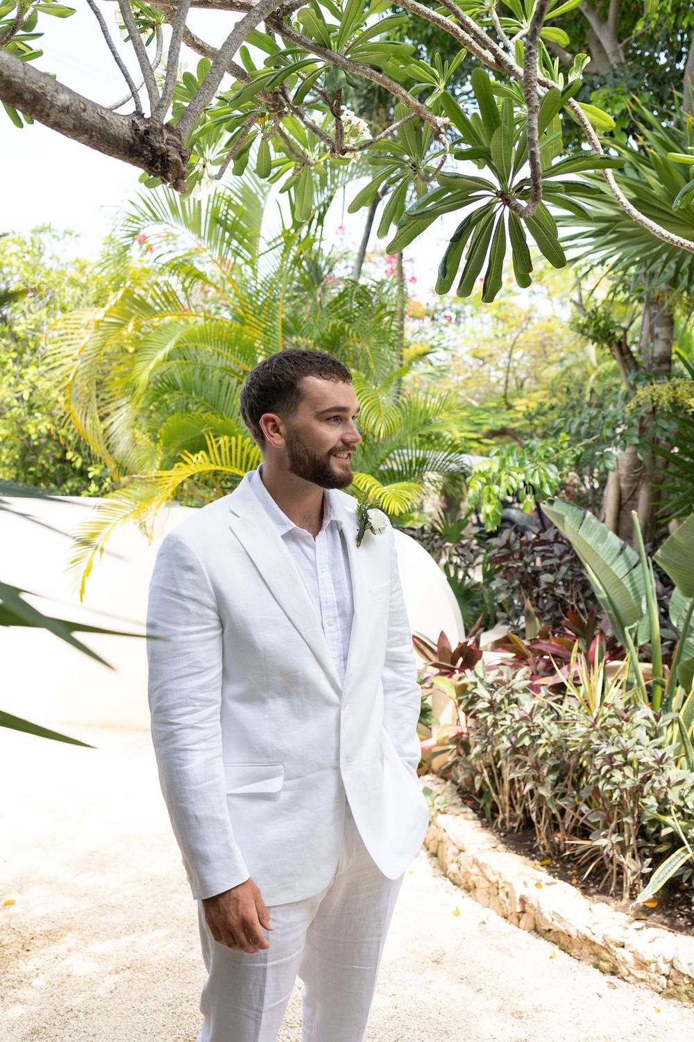 Groom in white linen suit smiling outdoors among tropical greenery on his wedding day.