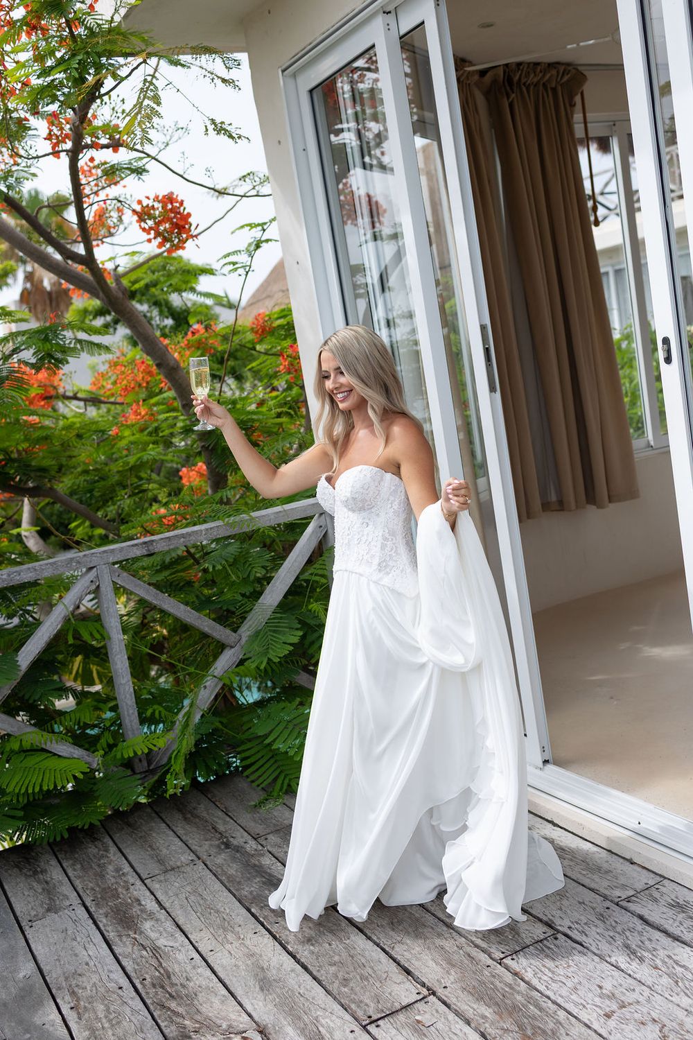 Radiant bride in strapless white gown holding champagne on a wooden deck surrounded by tropical blooms.