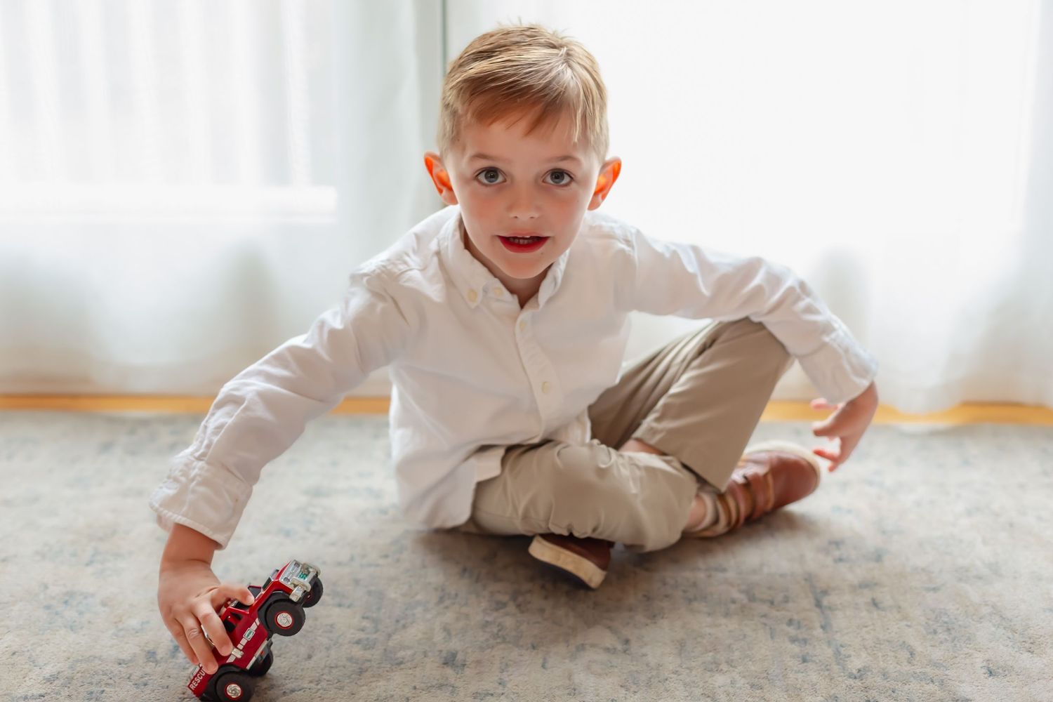Young boy in white shirt playing with red toy car on carpet floor near bright window.