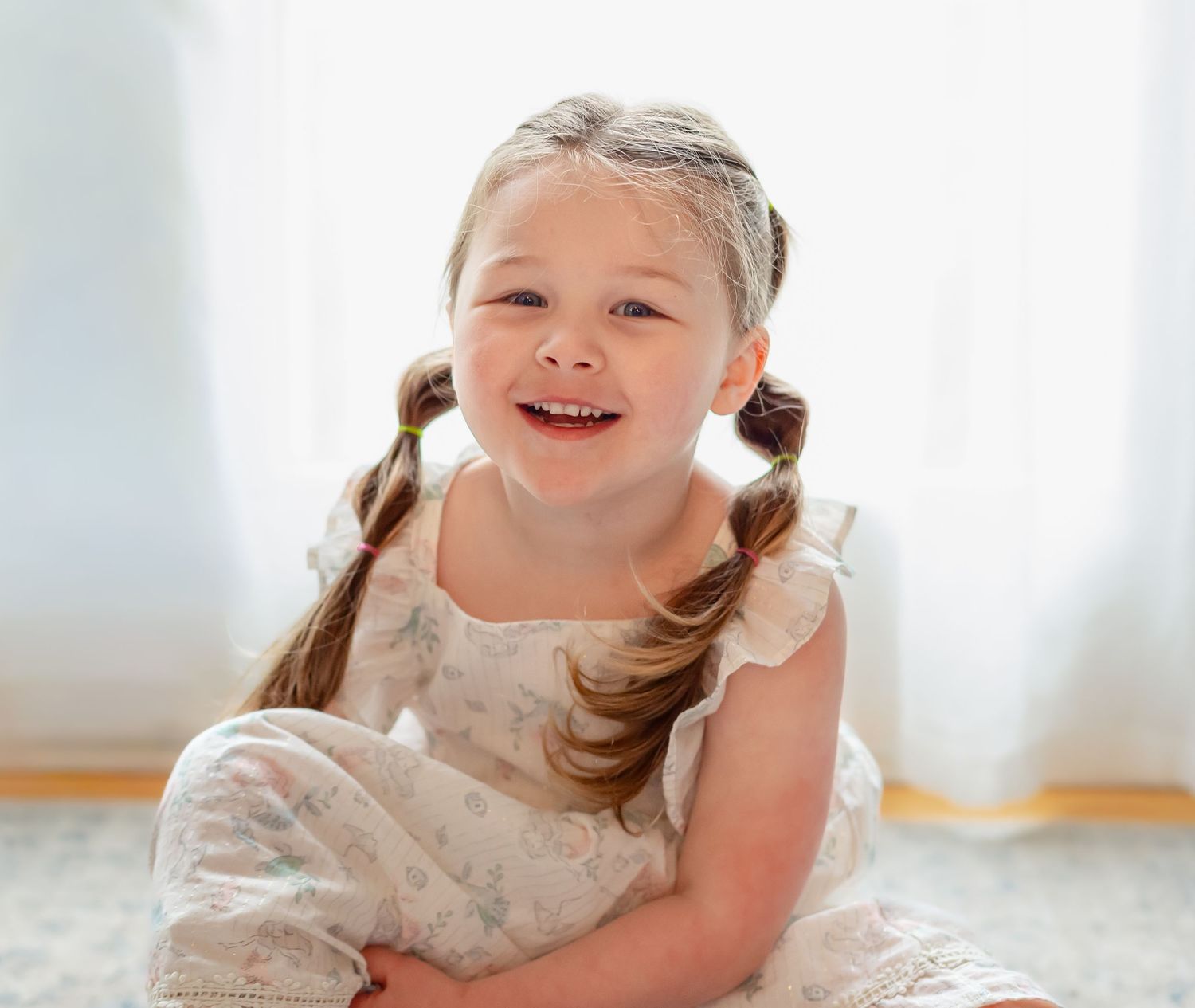 Smiling young girl with pigtails wearing a floral outfit, sitting on the floor in a bright, airy room.