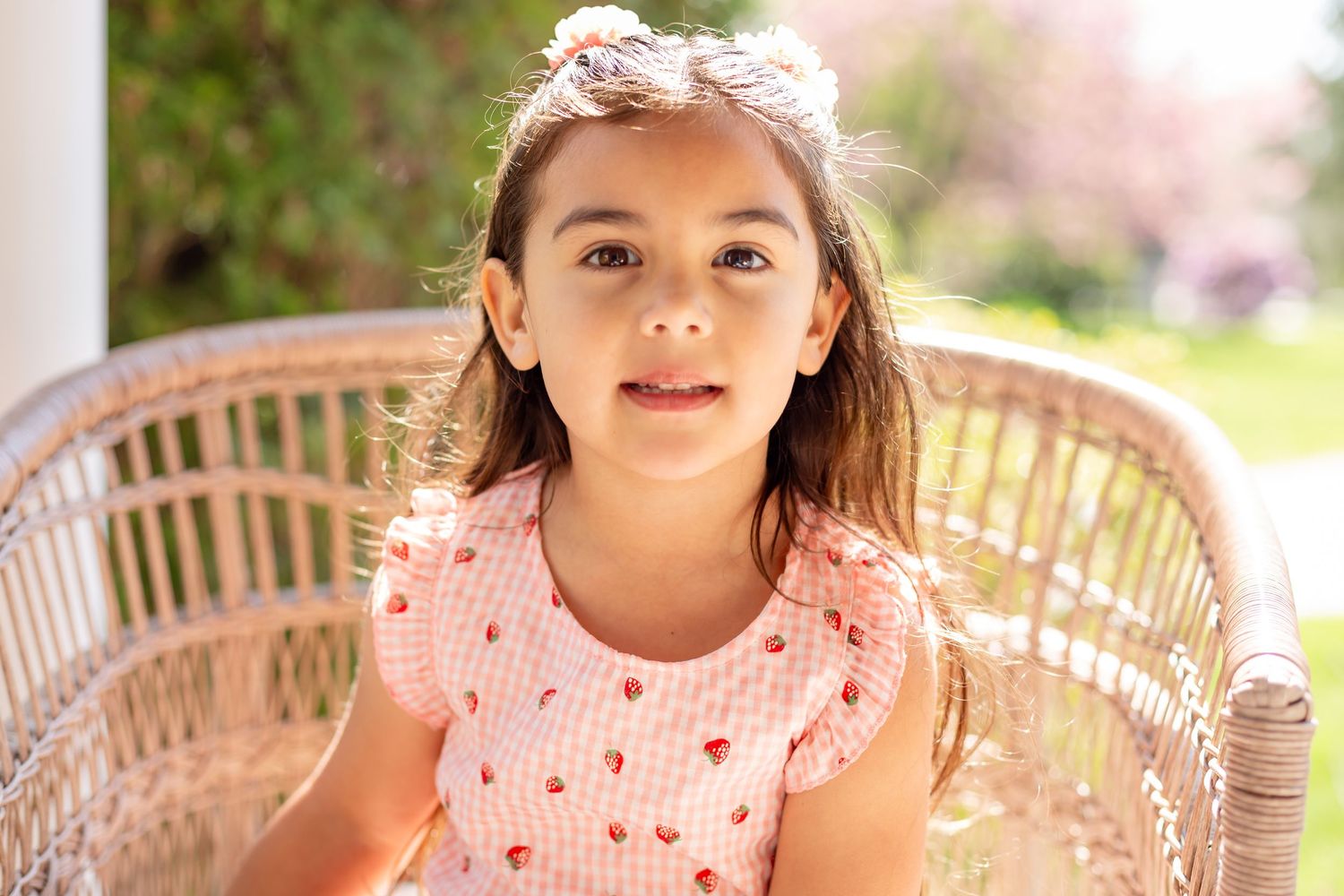 Young girl in pink gingham dress sitting in wicker chair outdoors with floral hair accessory.