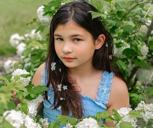 Young girl with dark hair surrounded by white blooming flowers and green leaves outdoors.