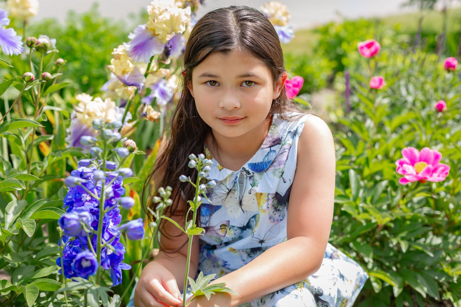 Young girl in floral dress crouching among vibrant garden flowers including blue delphiniums and pink blooms.