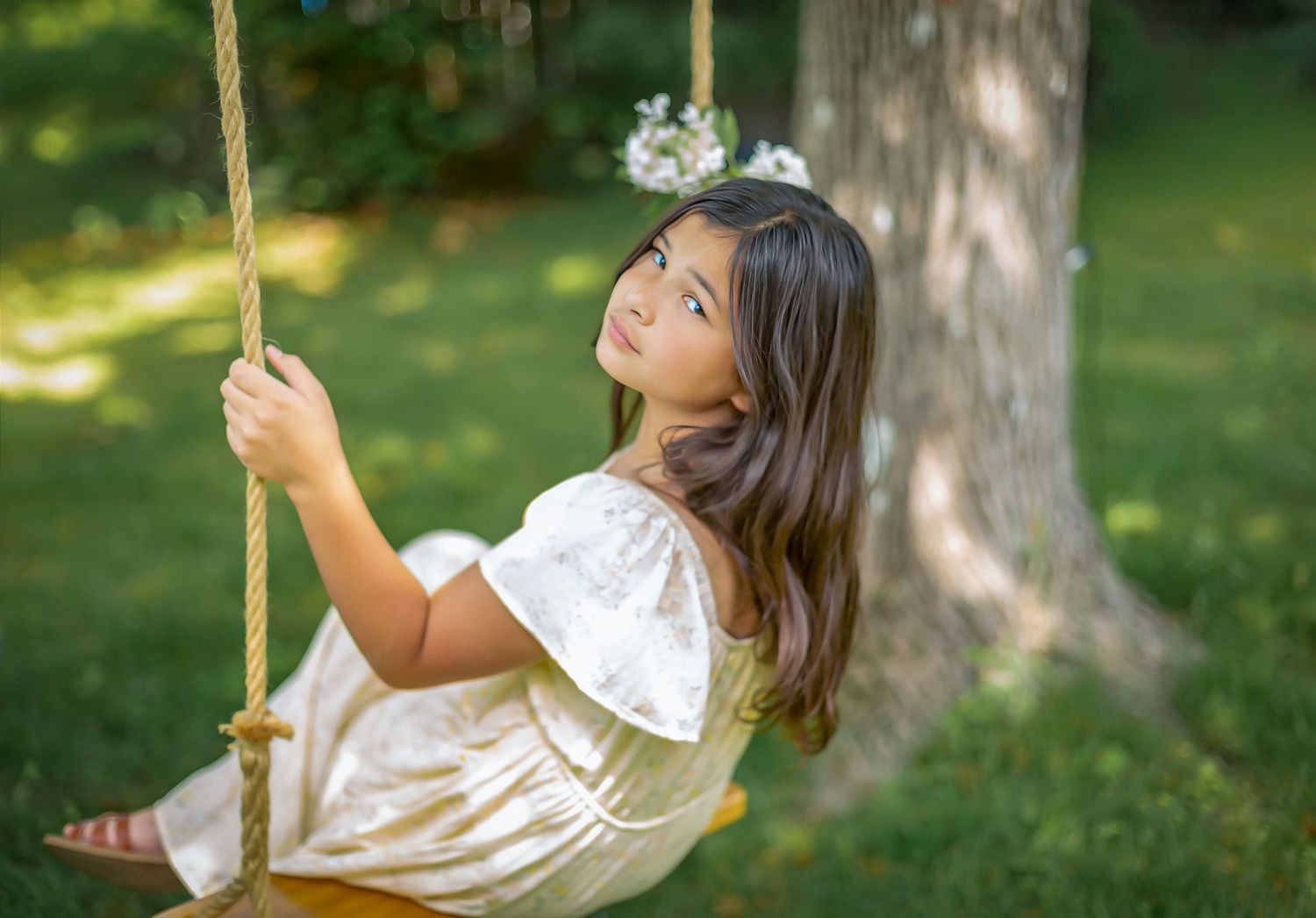 Young girl in white dress sitting on rope swing tied to tree, smiling outdoors in lush green garden.
