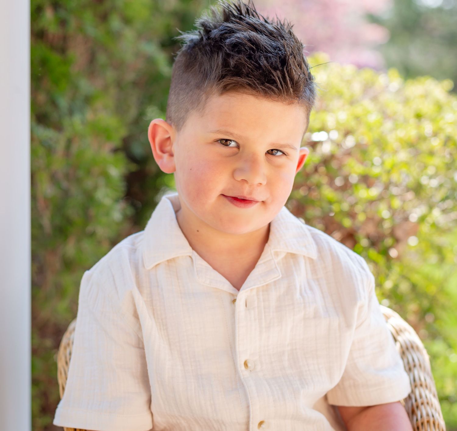 Young boy with spiked hair wearing a white linen shirt, sitting outdoors with green foliage in the background.
