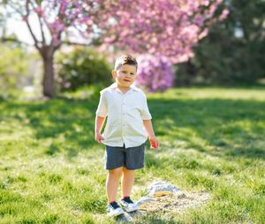 A young boy in a white shirt and blue shorts stands on grass with pink cherry blossom trees in the background.
