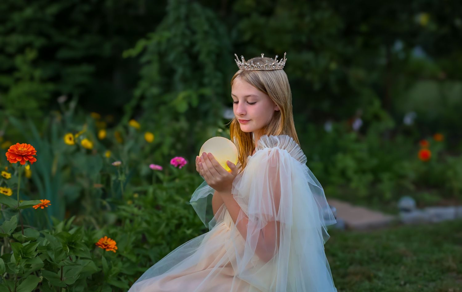 Young girl in princess crown and tulle dress holds glowing orb in lush garden at dusk.