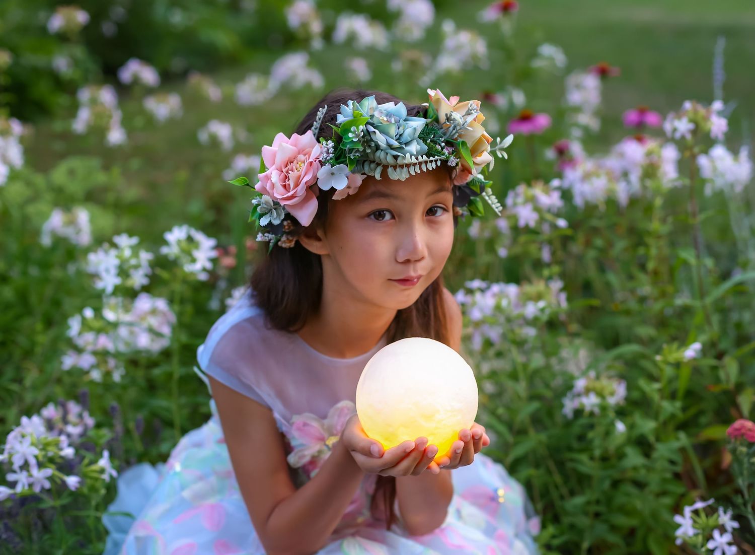 Young girl wearing floral crown holds glowing moon ball surrounded by blooming flowers in a garden.