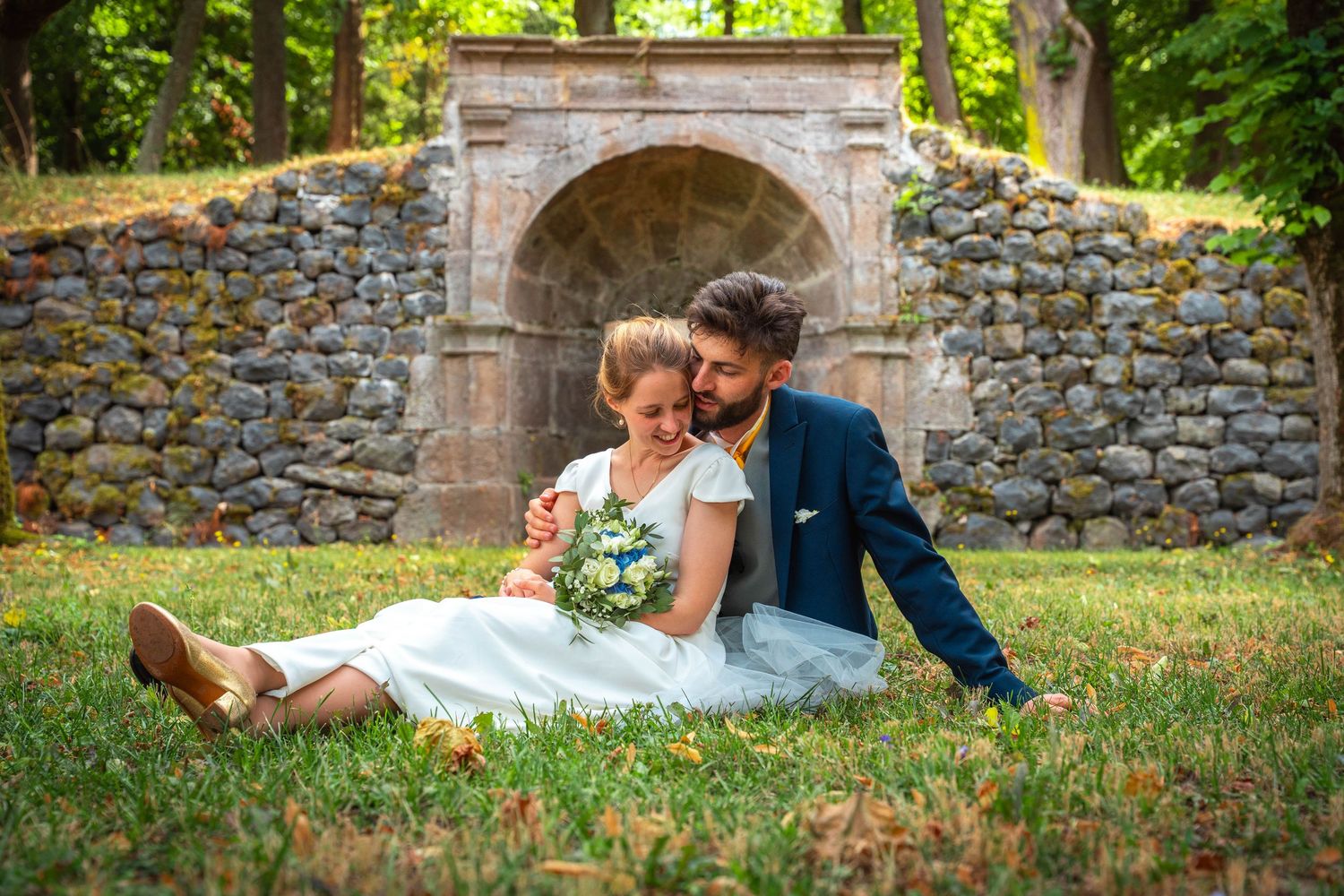 Newlywed couple sitting on grass in front of a stone arch, bride holding bouquet in a park setting.