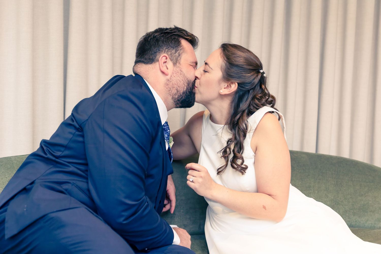 A bride in white dress and groom in navy suit share a tender kiss while seated on green couch.