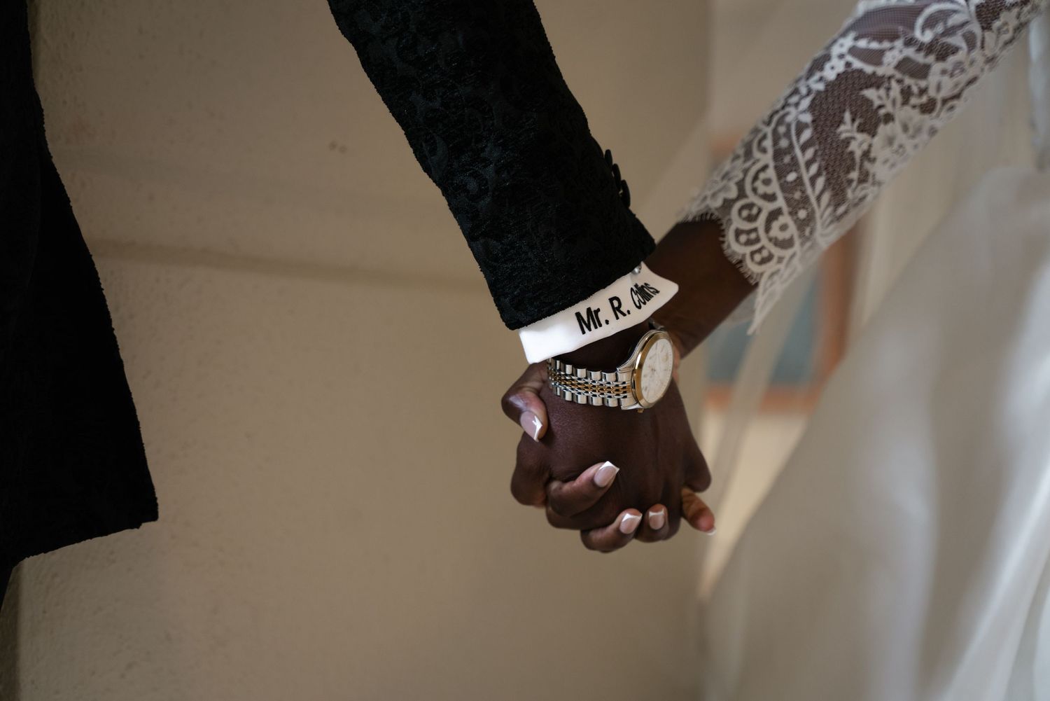 Close-up of interracial couple holding hands, groom wearing Mr. & Mrs. bracelet, bride in lace wedding dress.