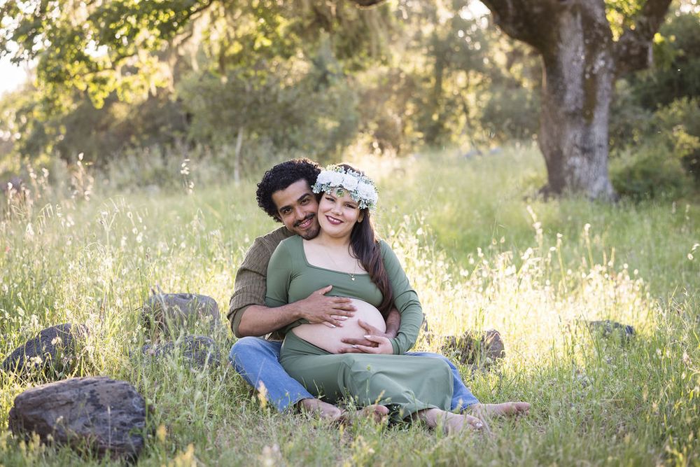 Couple sitting in a green meadow, man embracing pregnant woman wearing a floral crown.