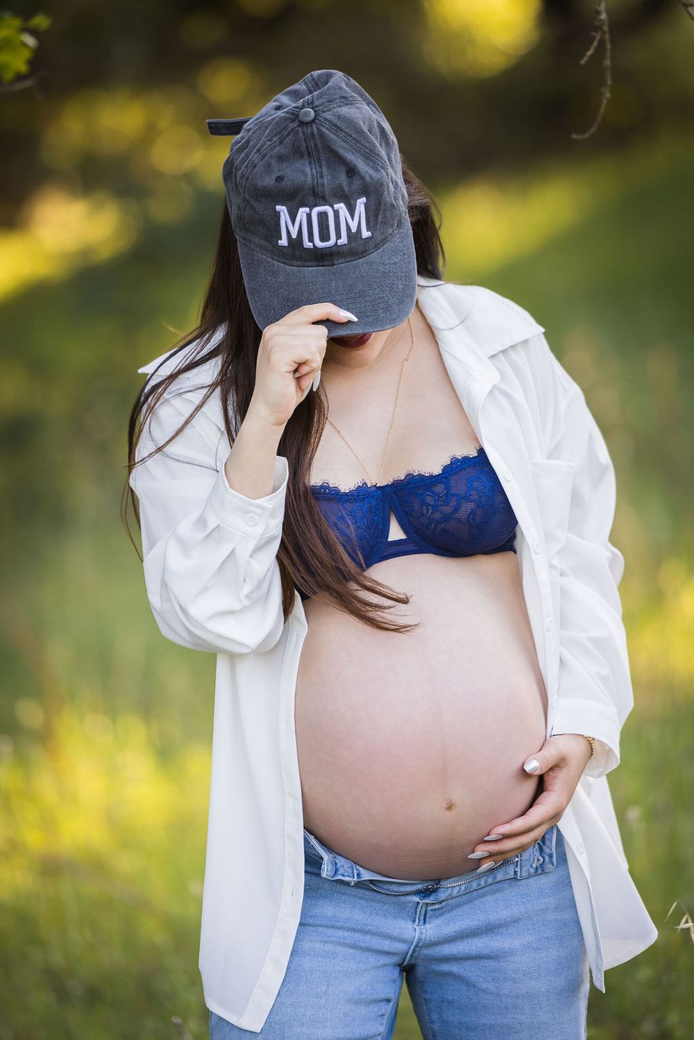 Pregnant woman in MOM cap, white shirt and blue lace bra cradles baby bump outdoors.