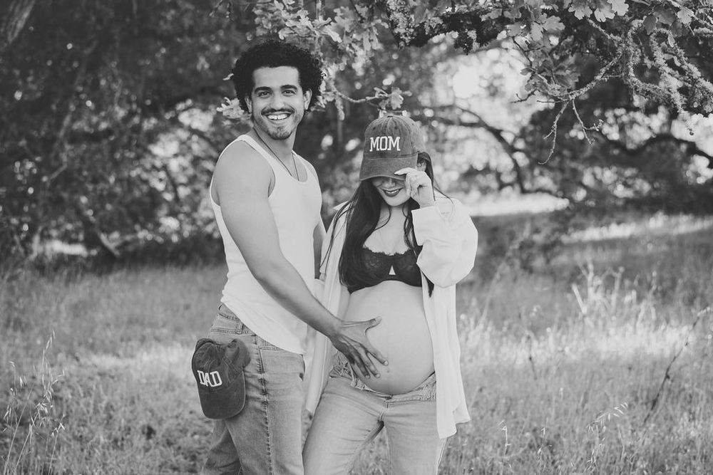 Black and white photo of smiling couple outdoors, woman wearing MOM cap, showing baby bump.
