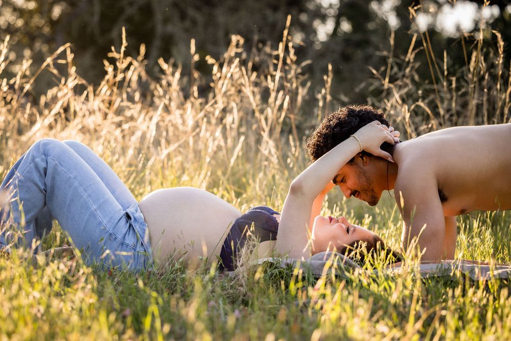 Shirtless man tenderly kissing pregnant woman's belly while lying in sunny grass field.