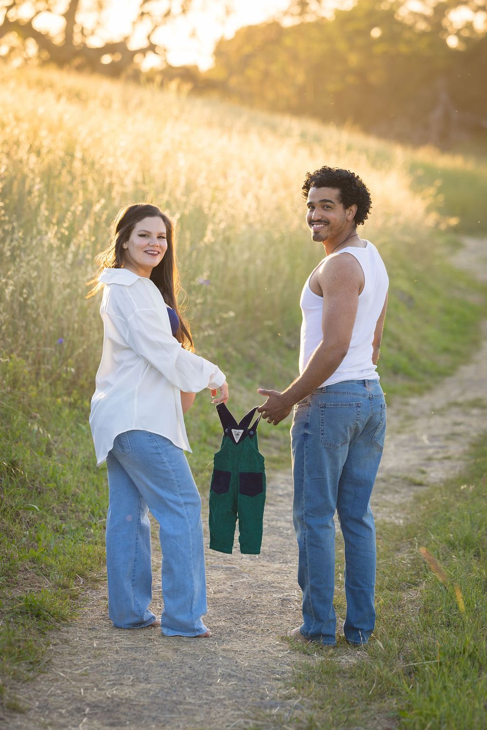 Expecting couple holding tiny green overalls on a sunlit dirt path surrounded by golden fields.
