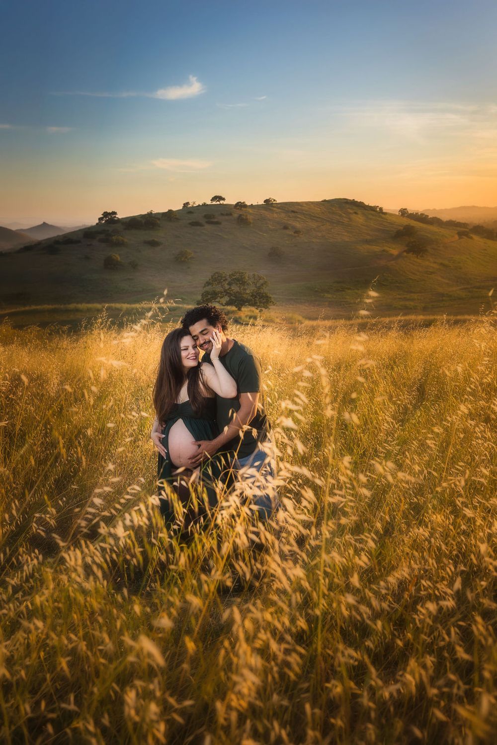 Couple sitting in golden field at sunset, man embracing pregnant woman with rolling hills in background.