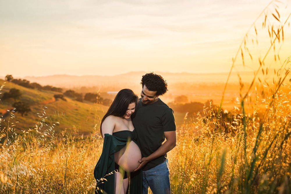 Pregnant couple embracing in a golden field at sunset with rolling hills in the background.