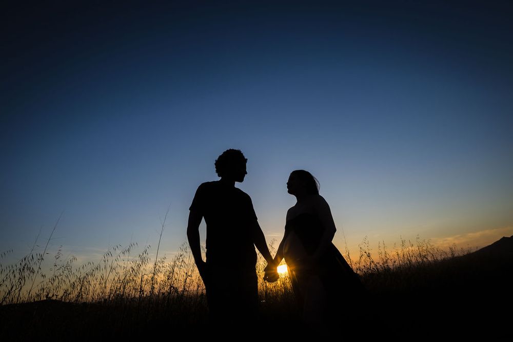 Silhouette of expecting couple holding hands against vibrant sunset sky in open field.