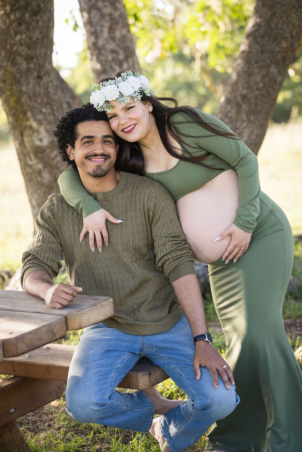 Smiling couple in matching green outfits pose outdoors for a maternity photo shoot under trees.