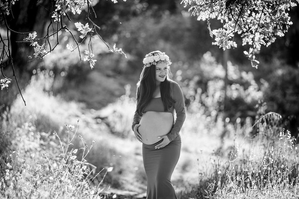 Black and white maternity photo of pregnant woman with flower crown in sunlit nature.