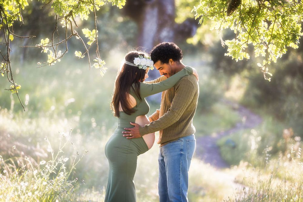 Couple embracing outdoors in golden sunlight, pregnant woman wearing flower crown in green dress.