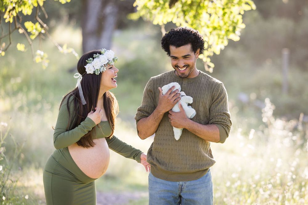 Happy pregnant couple in olive outerwear holding a white stuffed animal in a sunny meadow.