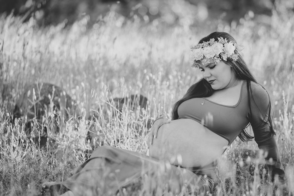 Black and white maternity photo of a pregnant woman wearing a floral crown sitting in a field of tall grass.
