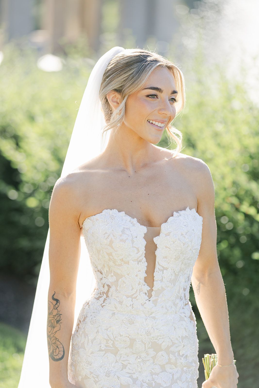 Armour House Wedding - Smiling bride in strapless floral lace wedding gown and veil, with arm tattoo, posing outdoors in soft sunlight.