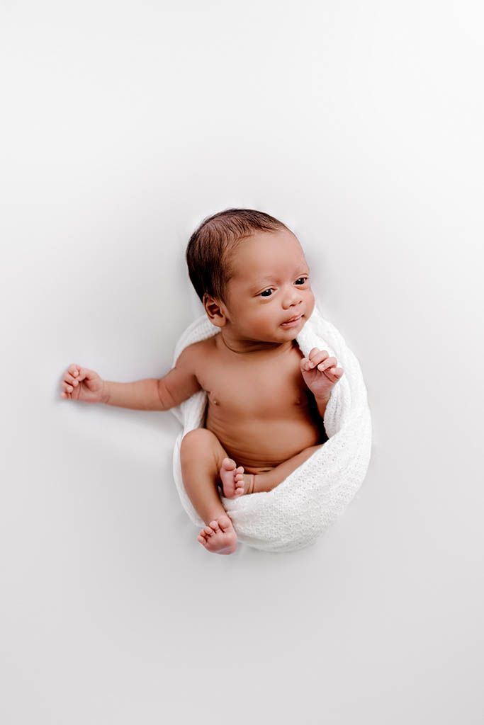 Newborn baby wrapped in white blanket lying on white background, alert with dark hair and tiny fists raised.