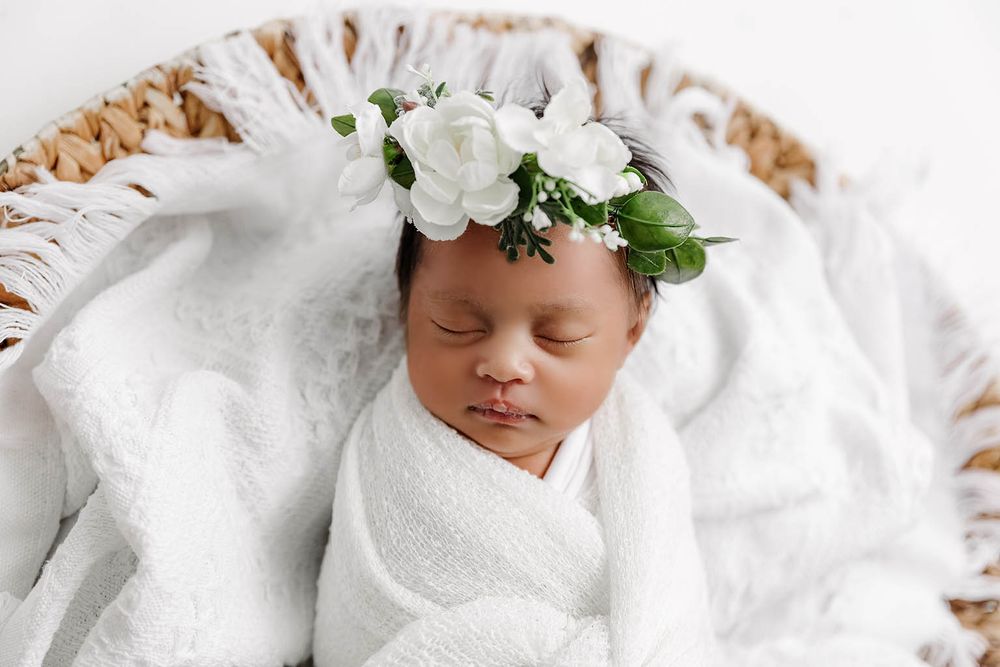 Sleeping newborn baby girl swaddled in white wrap with floral crown in wicker basket.