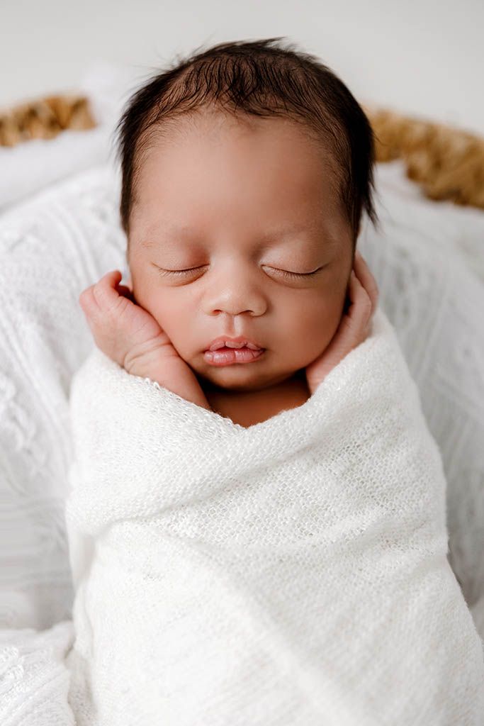 Peaceful newborn baby swaddled in white wrap sleeping in a basket with hands tucked under chin.