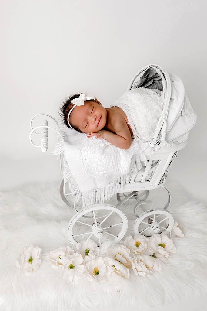 Sleeping newborn baby in white wrap and bow headband resting in a miniature white vintage pram with flowers.