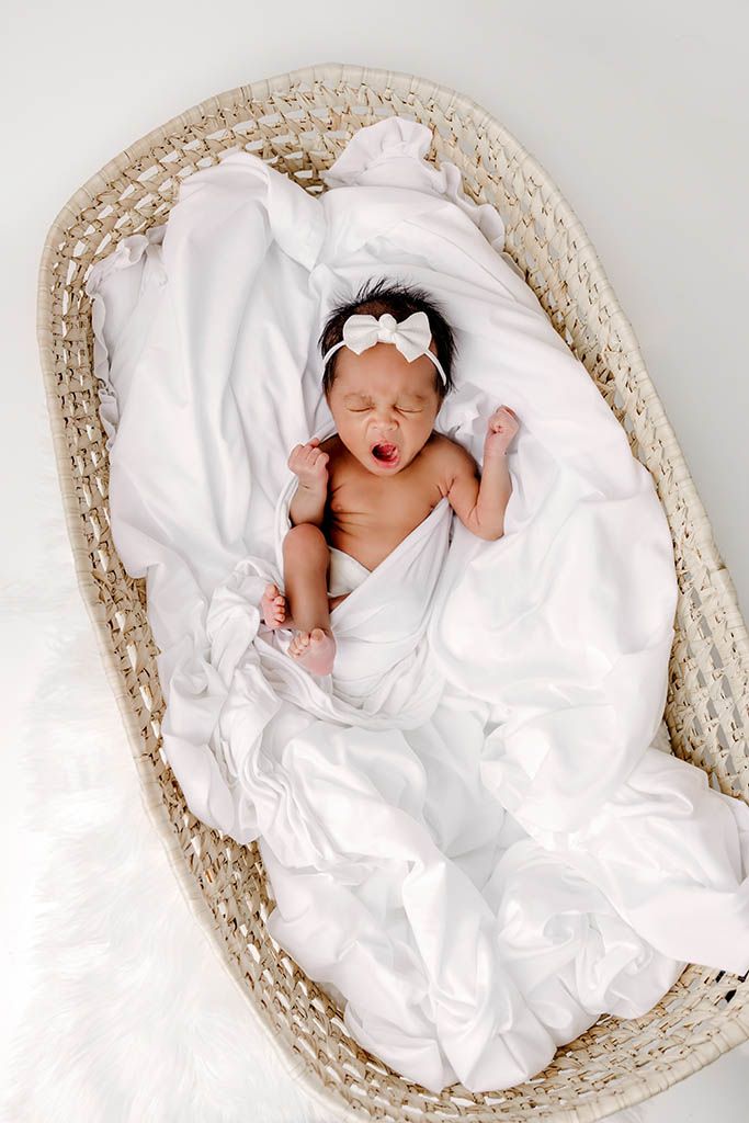 Newborn baby girl yawning in white wrap with bow headband lying in oval wicker bassinet.