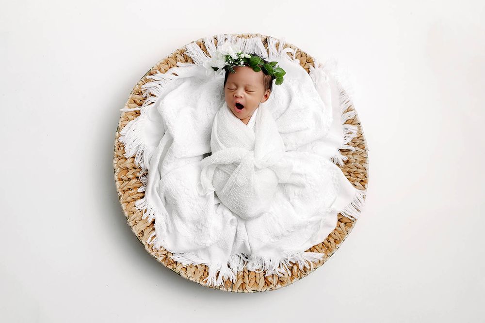 Newborn baby swaddled in white wrap wearing a green floral crown, sleeping in a wicker basket on white background.