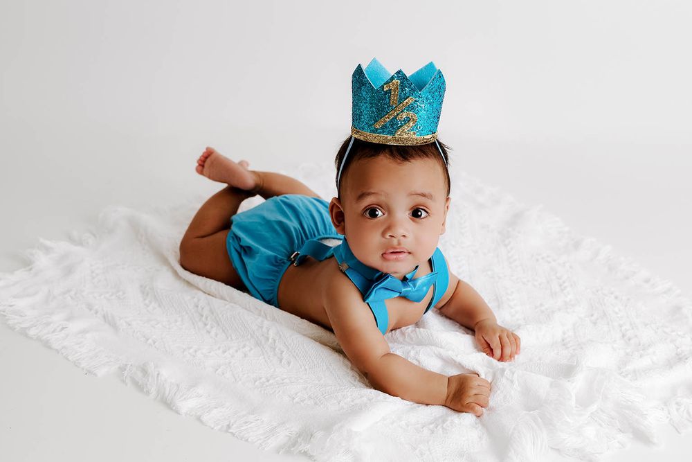 Adorable baby in teal outfit and gold crown lying on white blanket.