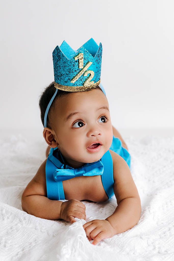 Baby in blue bow tie and glittery crown celebrating half birthday milestone, lying on white blanket.