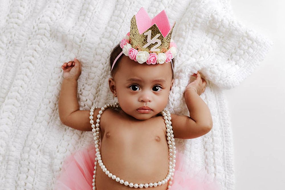Baby girl in pink tutu and pearl necklace wearing gold glitter half birthday crown on white knit blanket.