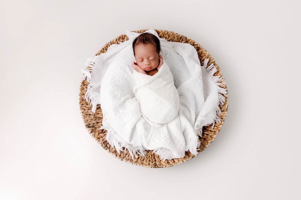 Newborn baby swaddled in white wrap sleeping peacefully in a round woven basket on white background.