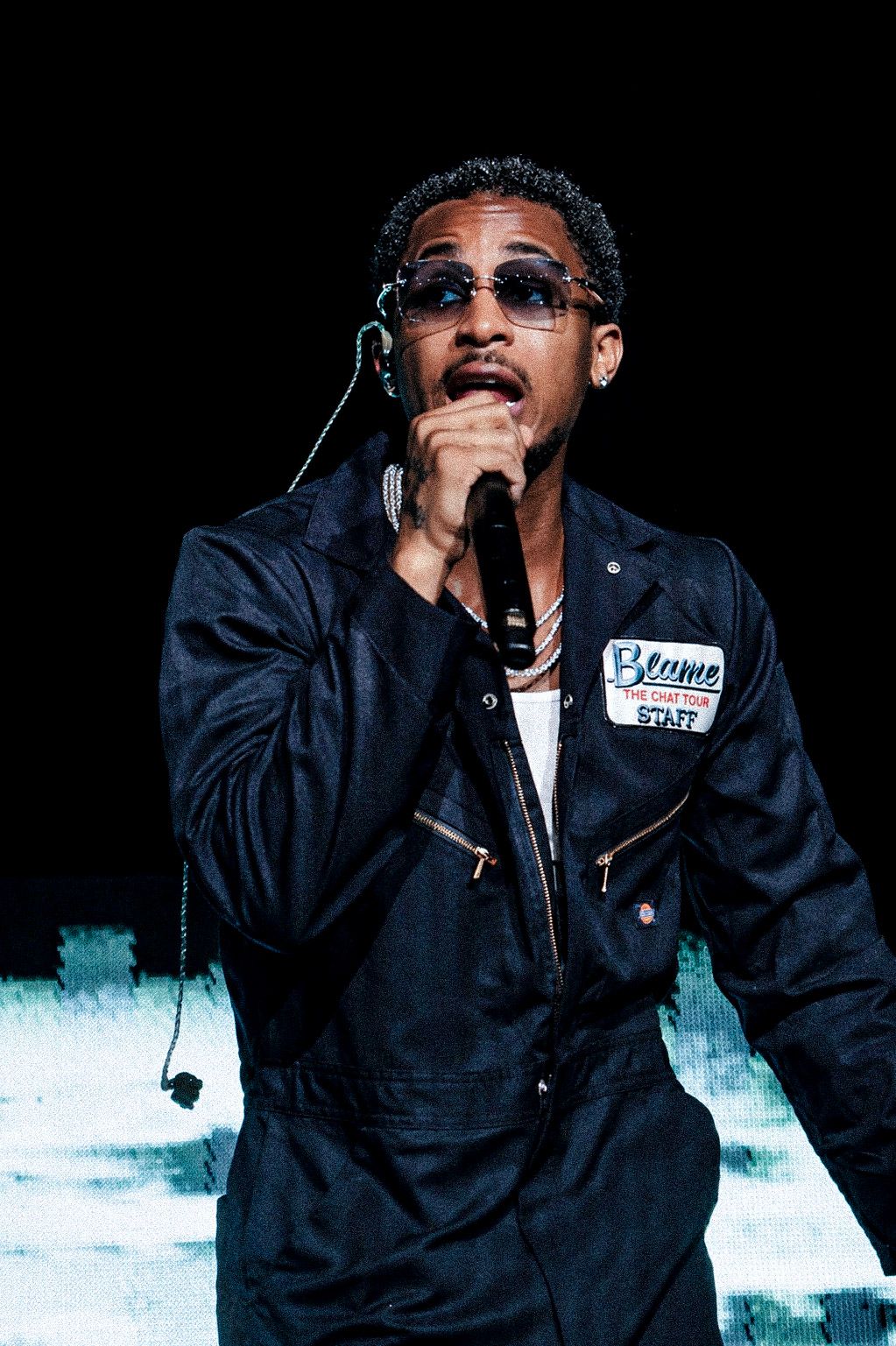 Male performer singing into microphone on stage, wearing dark denim jumpsuit with staff patch and tinted sunglasses.