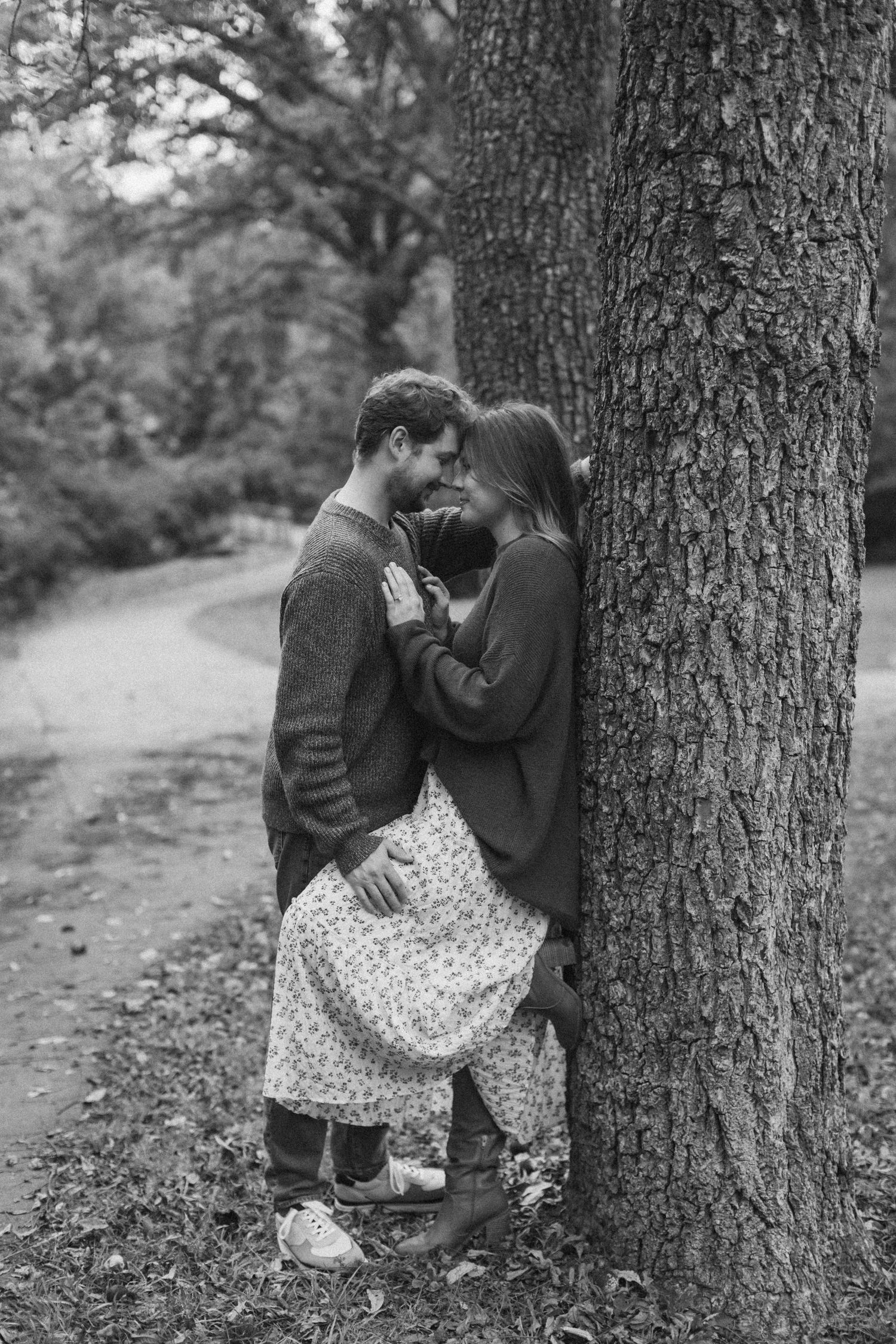 A woman stands with her back against a tree trunk, one foot propped up as her fiance leans in toward her, touching his forehead to hers.