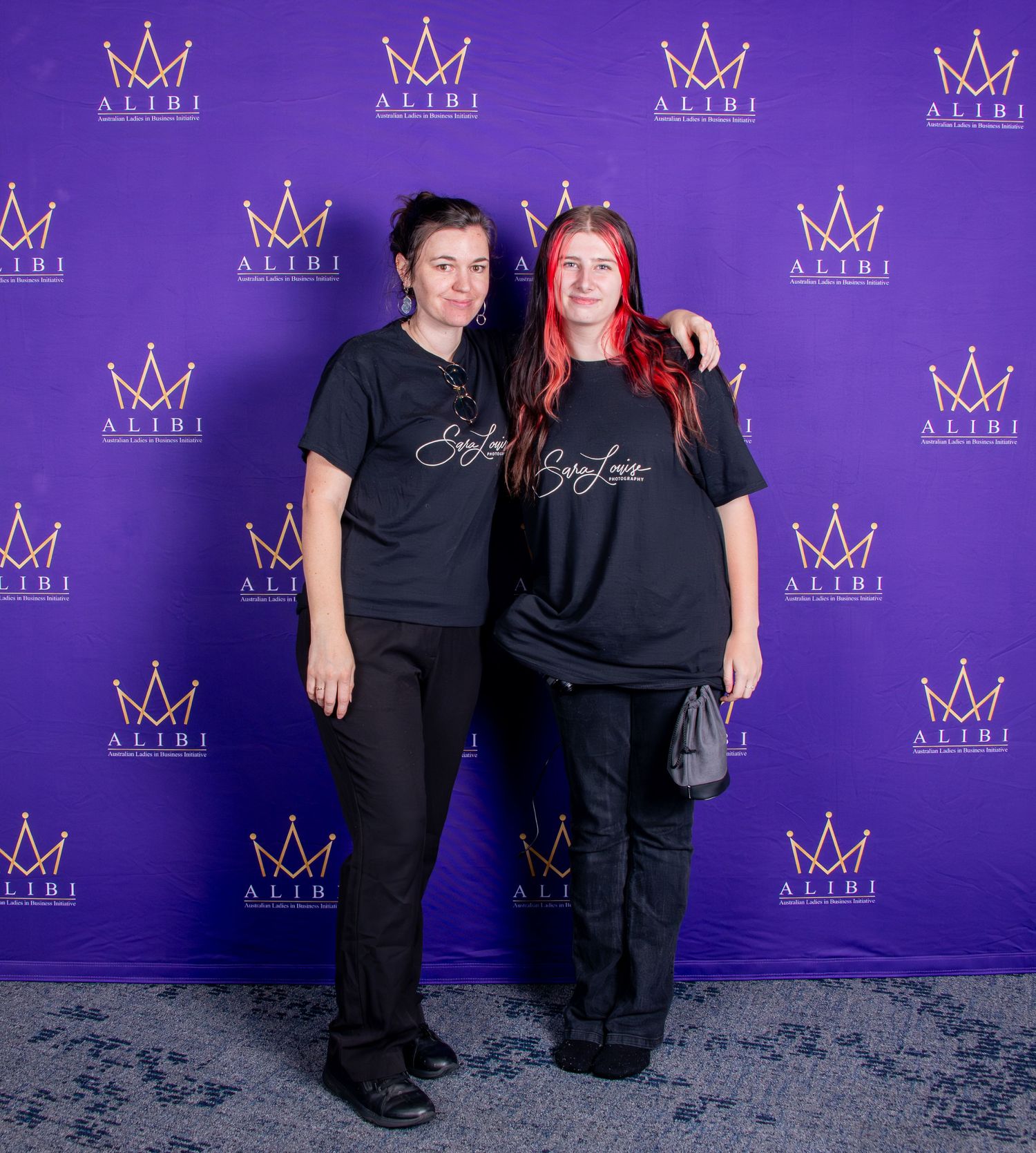Two women in black Alibi branded shirts posing in front of a purple Alibi logo backdrop.