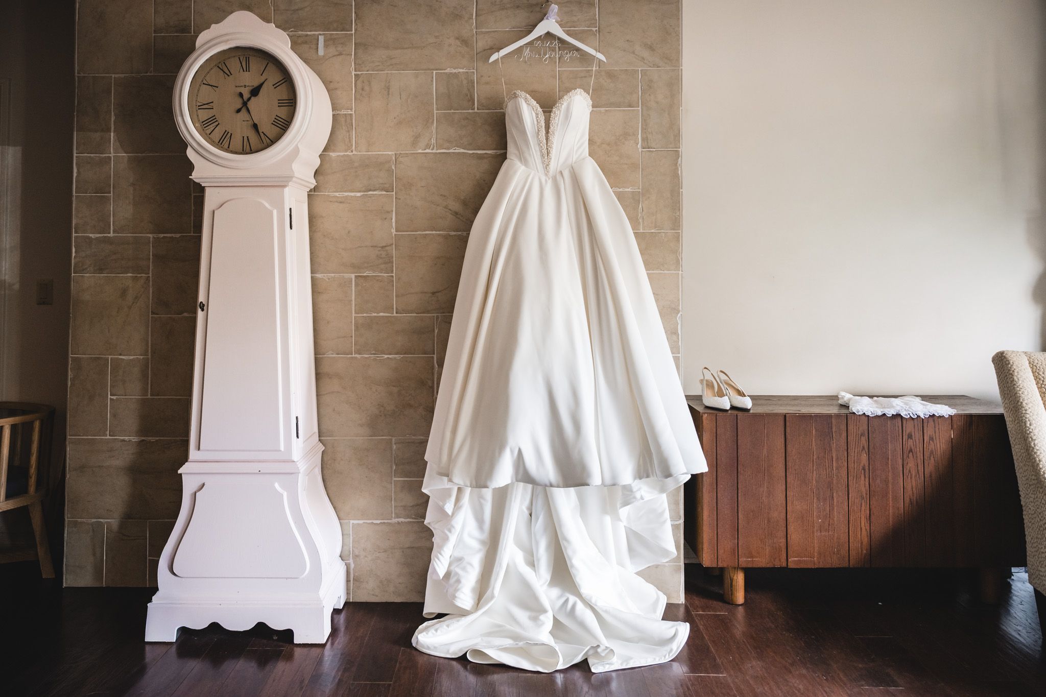 Elegant strapless wedding gown hanging on wall beside white grandfather clock and wooden dresser.
