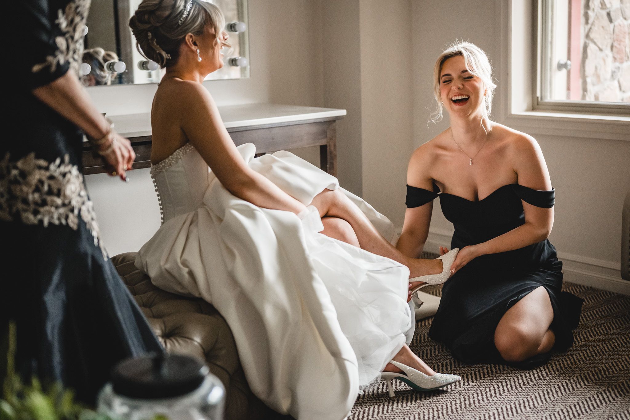 Bridesmaid in black off-shoulder dress helps bride with shoes while laughing during wedding preparations.