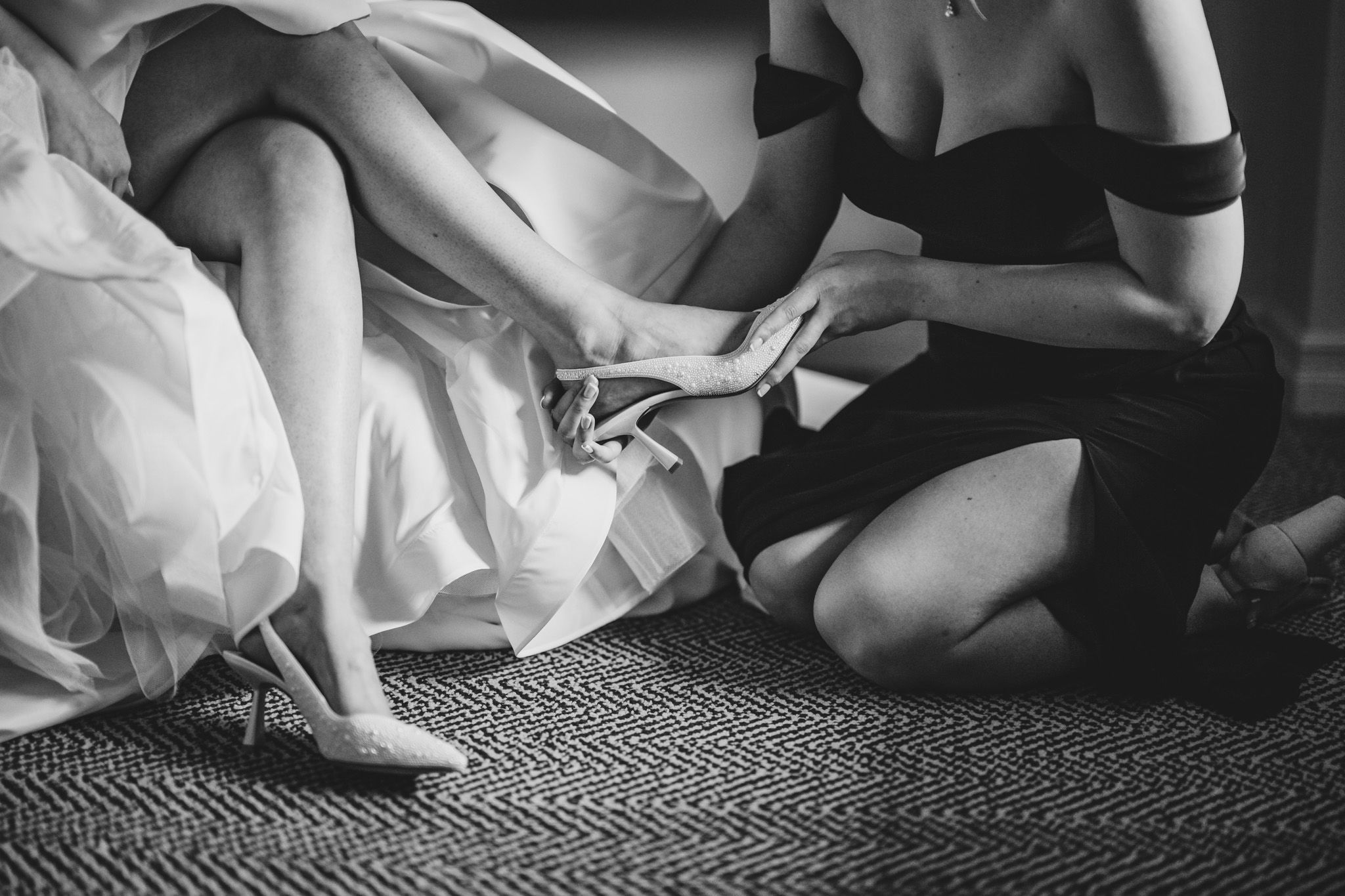 Black and white photo of bridesmaid helping bride put on pearl wedding heels before the ceremony.